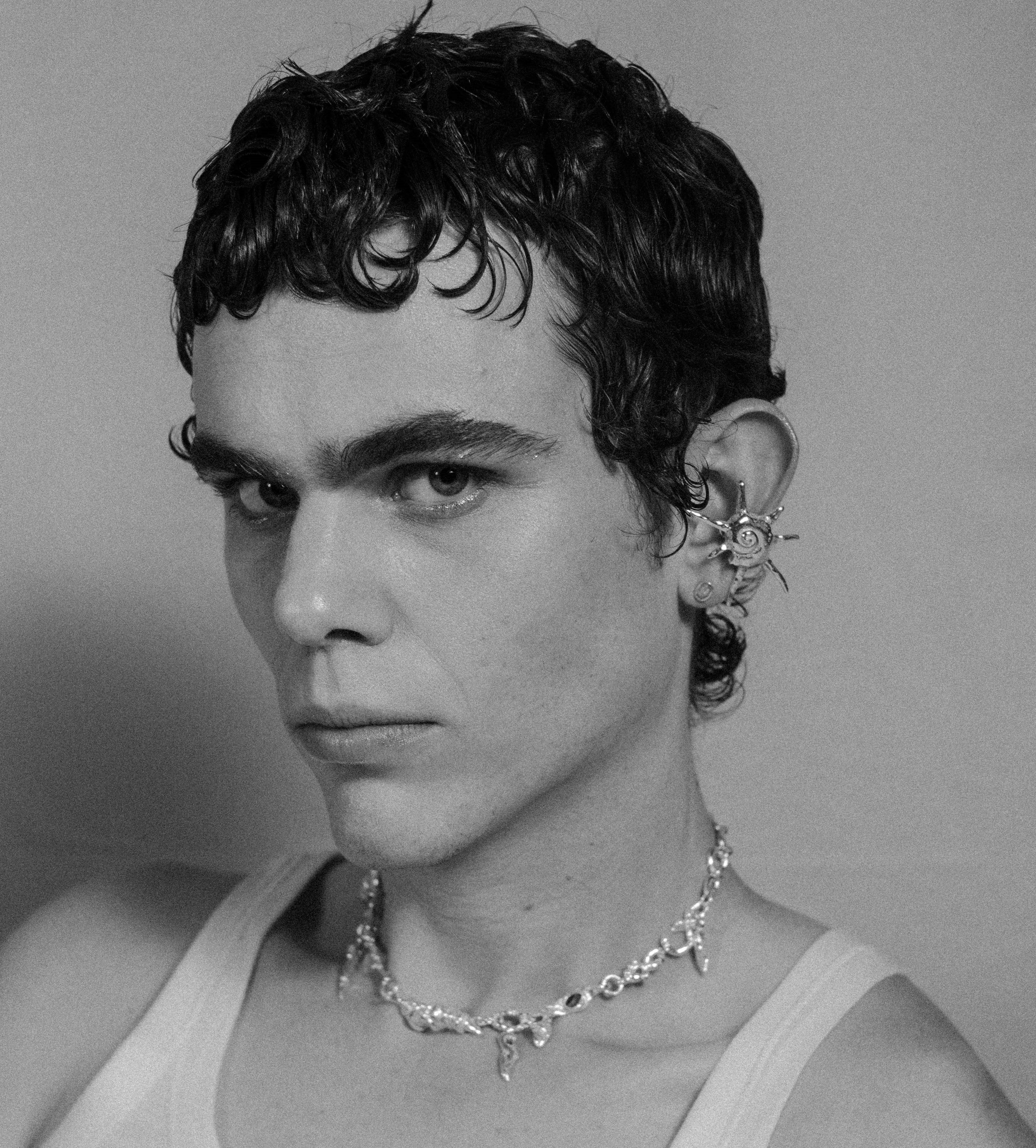 Young man in white tank top with rings on his fingers, his palms holding his face in a 3/4 position, looking straight to the camera